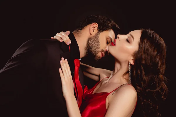 Vista lateral del hombre guapo besando elegante novia con labios rojos aislados en negro - foto de stock