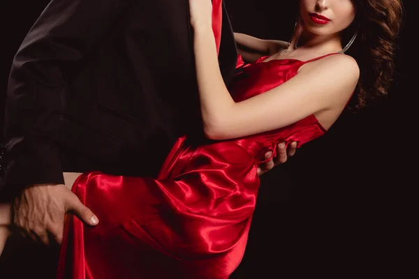 Vista recortada de hombre elegante abrazando y sosteniendo por mujer seductora led en vestido rojo aislado en negro - foto de stock