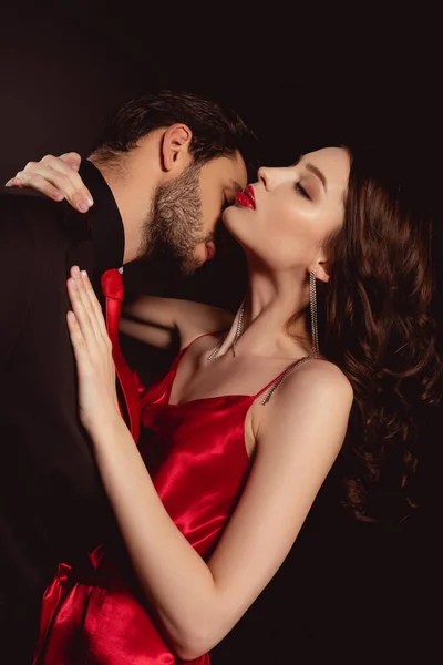 Vista laterale di uomo in giacca e cravatta baciare donna attraente in abito rosso isolato su nero — Foto stock