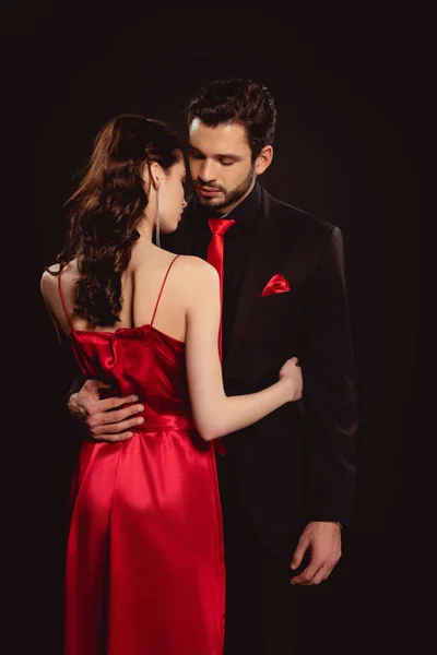 Hombre guapo en traje abrazando por la cintura novia elegante en vestido rojo aislado en negro - foto de stock