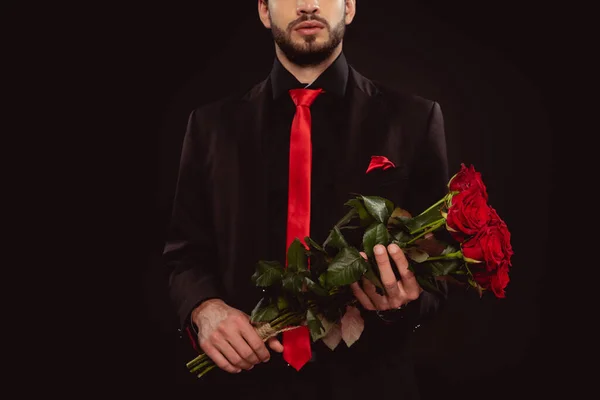 Обрезанный вид человека в костюме с красными розами, изолированными на черном — стоковое фото