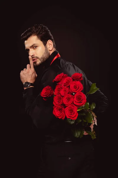 Vue latérale du bel homme tenant un bouquet de roses et montrant un signe secret isolé sur noir — Photo de stock