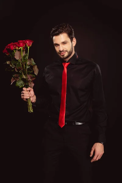 Hombre guapo en ropa formal y corbata roja mirando a la cámara y sosteniendo ramo de rosas aisladas en negro - foto de stock