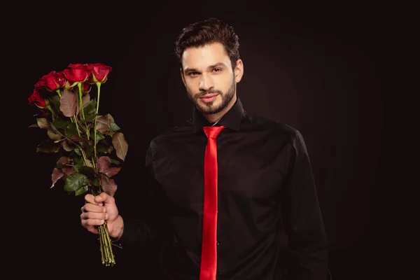 Homem elegante olhando para a câmera e segurando buquê de rosas vermelhas isoladas em preto — Fotografia de Stock