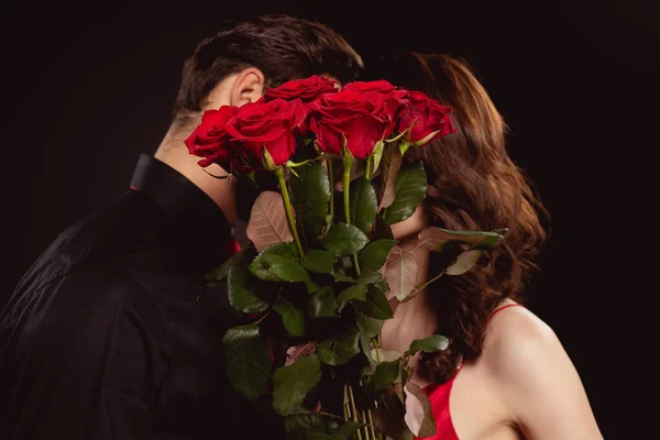 Vista lateral de la elegante pareja cubriendo caras con rosas rojas aisladas en negro - foto de stock