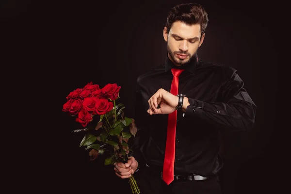 Handsome man looking at wristwatch and holding bouquet of red roses isolated on black — Stock Photo