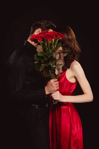 Vue latérale du couple élégant couvrant les visages avec bouquet de roses rouges isolé sur noir — Photo de stock