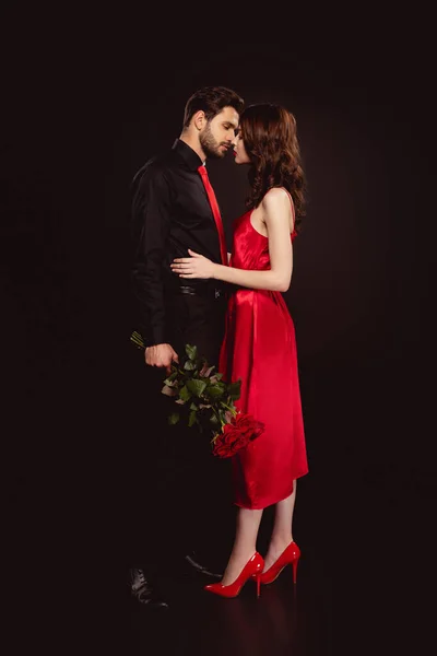 Vista laterale dell'uomo in abbigliamento formale tenendo bouquet di rose rosse e baciare donna in abito isolato su nero — Foto stock