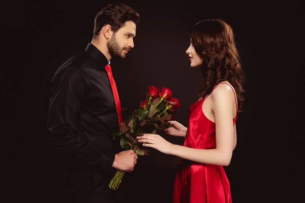 Vista lateral del hombre guapo dando rosas a novia atractiva aislada en negro - foto de stock
