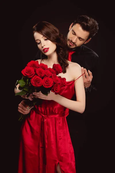 Handsome man taking off dress from elegant girlfriend with red roses isolated on black — Stock Photo