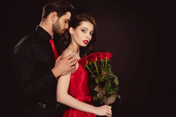 Vista lateral del hombre elegante abrazando novia con rosas rojas aisladas en negro - foto de stock