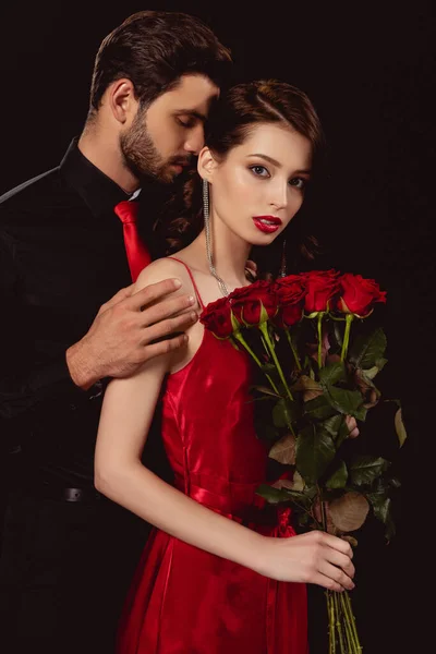 Homme élégant embrassant femme attrayante avec bouquet de roses rouges isolé sur noir — Photo de stock