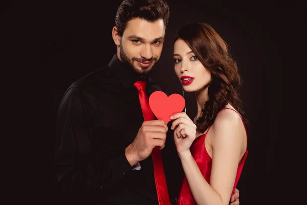 Elegant couple holding paper heart and looking at camera isolated on black — Stock Photo