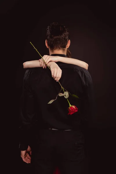 Vista posterior de la mujer sosteniendo rosa y abrazando novio aislado en negro - foto de stock