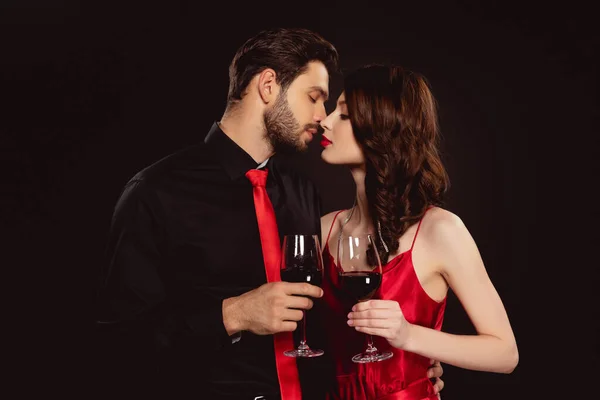 Vue latérale de couple élégant baisers et vin rouge clinquant isolé sur noir — Photo de stock