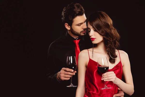 Elegante hombre abrazando hermosa novia con copa de vino tinto aislado en negro - foto de stock