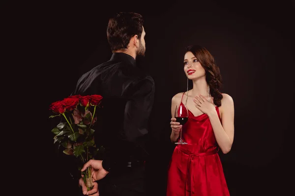 Homme élégant cachant bouquet de roses près de petite amie souriante avec du vin rouge isolé sur noir — Photo de stock