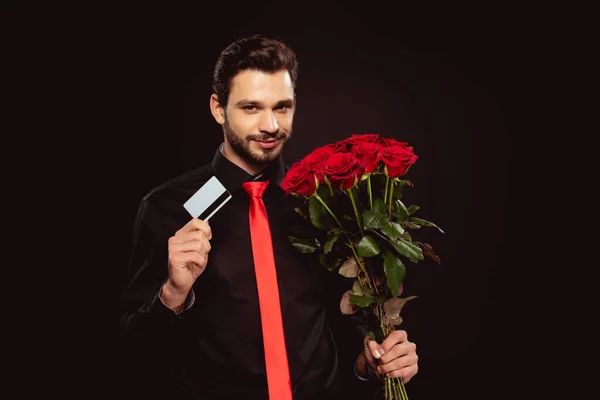 Handsome man holding credit card and roses while looking at camera isolated on black — Stock Photo