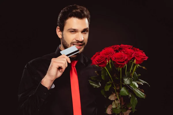 Handsome man biting credit card and holding bouquet of roses isolated on black — Stock Photo