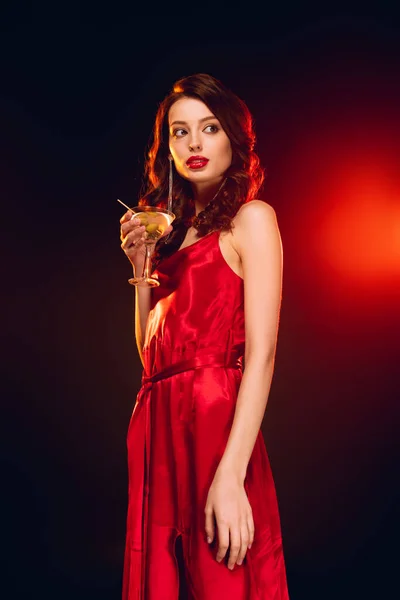 Elegant girl in red dress holding glass of martini on black background with lighting — Stock Photo