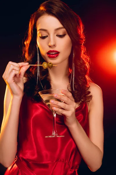 Hermosa mujer sosteniendo palo con aceitunas y vaso de martini sobre fondo negro con iluminación - foto de stock