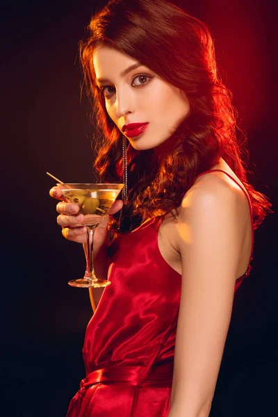 Vista lateral de una hermosa mujer en vestido sosteniendo un vaso de martini y mirando a la cámara sobre fondo negro con iluminación - foto de stock