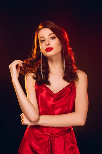 Attractive girl in red dress touching hair and looking at camera isolated on black — Stock Photo