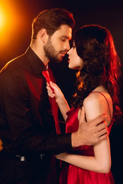 Vista lateral homem elegante beijando bela namorada em vestido vermelho no fundo preto com iluminação — Fotografia de Stock