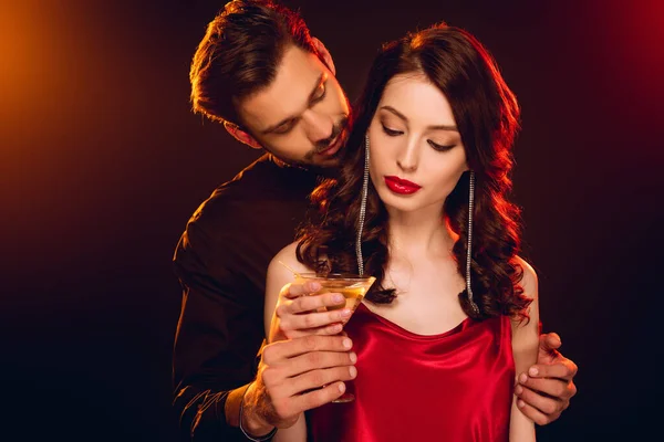 Hombre guapo abrazando novia elegante y sosteniendo vaso de cóctel sobre fondo negro con iluminación - foto de stock