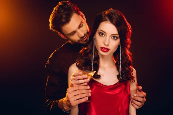 Elegante donna in abito rosso in possesso di vetro di cocktail vicino bel fidanzato su sfondo nero con illuminazione — Foto stock