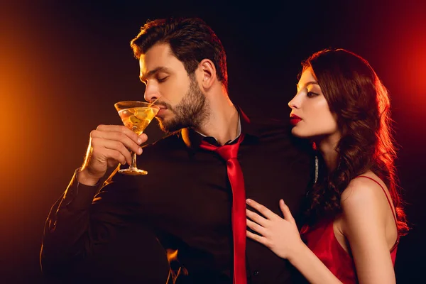 Schöner Mann trinkt Cocktail in der Nähe eleganter Freundin auf schwarzem Hintergrund mit Beleuchtung — Stockfoto