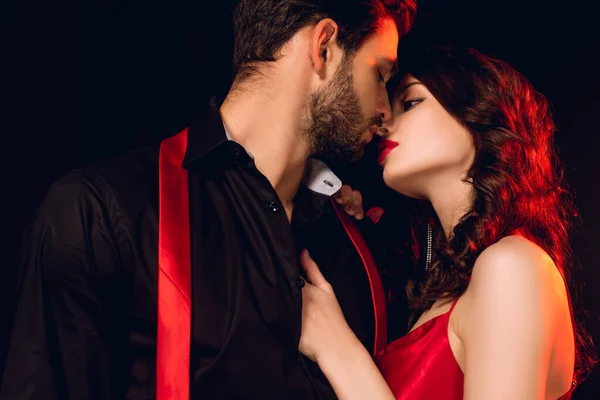 Hermosa mujer con labios rojos besando novio guapo con corbata desatada aislado en negro - foto de stock