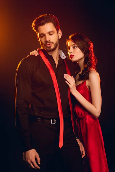 Attractive girl in red dress hugging handsome boyfriend with untied tie on black background with lighting — Stock Photo