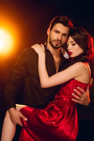 Handsome man looking at camera while hugging beautiful girlfriend in red dress on black background with lighting — Stock Photo