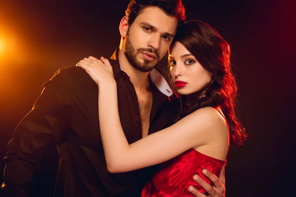 Hermosa mujer en vestido rojo mirando a la cámara mientras abraza hombre guapo sobre fondo negro con iluminación - foto de stock