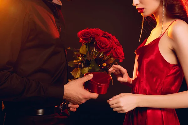 Vista recortada del hombre dando caja de regalo y ramo de rosas a la novia elegante sobre fondo negro con iluminación - foto de stock