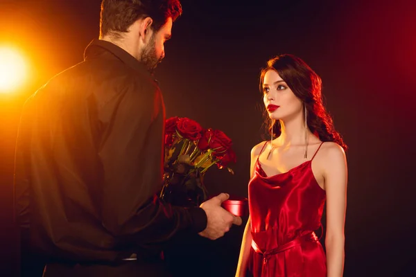 Mann mit Geschenkschachtel und Rosen in der Nähe einer eleganten Freundin im roten Kleid auf schwarzem Hintergrund mit Beleuchtung — Stockfoto