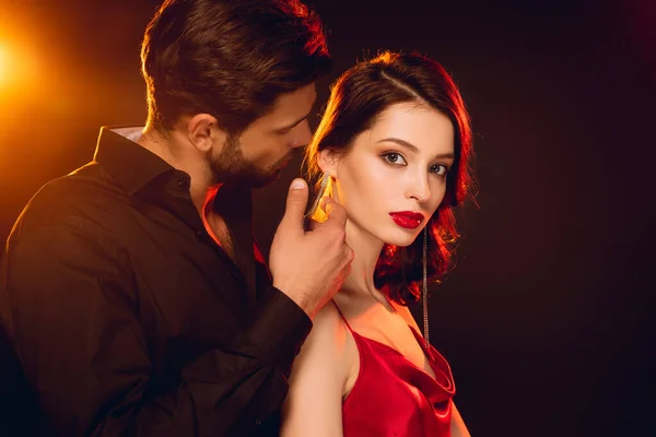 Side view of handsome man touching neck of beautiful girlfriend looking at camera on black background with lighting — Stock Photo