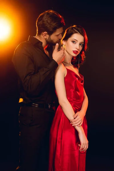 Вид сбоку элегантного мужчины, трогающего шею привлекательной подруги в красном платье на черном фоне с подсветкой — стоковое фото