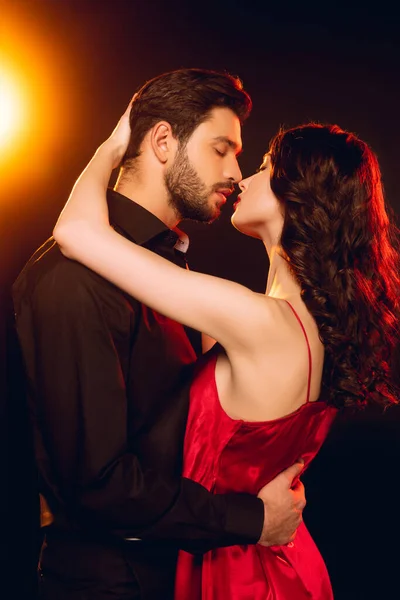 Vista lateral de mujer elegante en vestido rojo abrazando y besando novio guapo sobre fondo negro con iluminación - foto de stock