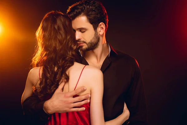 Handsome man embracing girlfriend in red dress on black background with lighting — Stock Photo