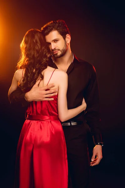 Elegante hombre mirando a la cámara mientras abraza a su novia en vestido rojo sobre fondo negro con iluminación - foto de stock