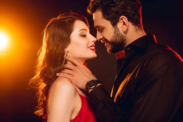 Side view of handsome man touching neck of smiling girlfriend in red dress on black background with lighting — Stock Photo