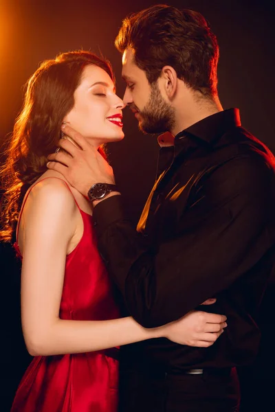 Side view of elegant man touching neck of beautiful smiling girl on black background with lighting — Stock Photo