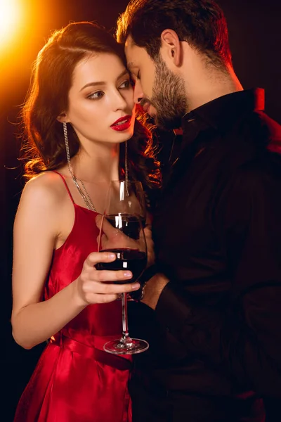 Mulher bonita no vestido segurando copo de vinho perto de namorado bonito no fundo preto com iluminação — Fotografia de Stock