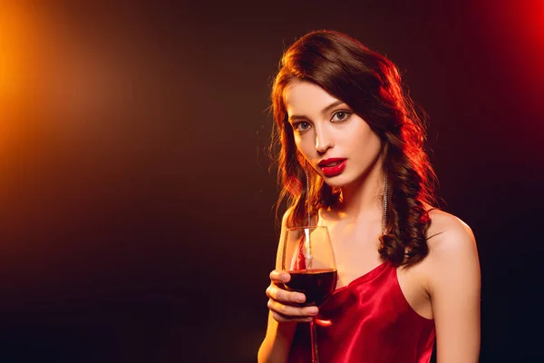 Елегантна дівчина в червоній сукні тримає келих вина і дивиться на камеру на чорному тлі з освітленням — стокове фото