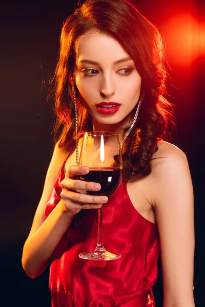 Schöne Frau im roten Kleid hält ein Glas Wein auf schwarzem Hintergrund mit Beleuchtung — Stockfoto