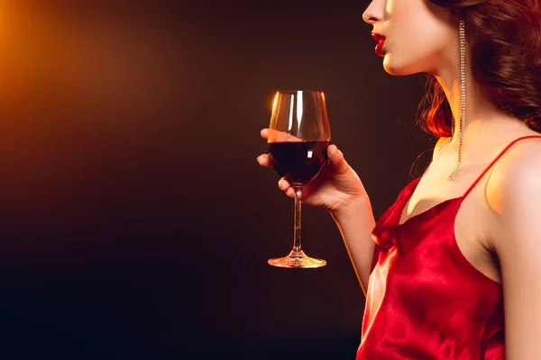 Cropped view of elegant woman with red lips holding glass of wine on black background with lighting — Stock Photo