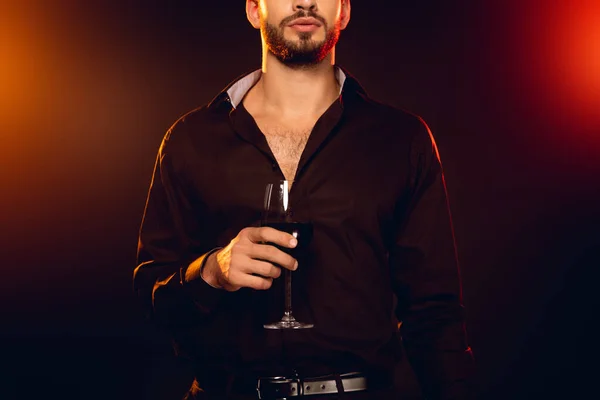 Cropped view of elegant man holding glass of red wine on black background with lighting — Stock Photo