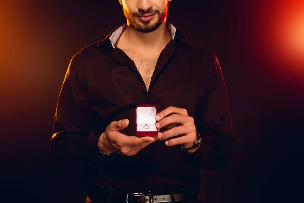 Cropped view of elegant man holding gift box with jewelry ring on black background with lighting — Stock Photo